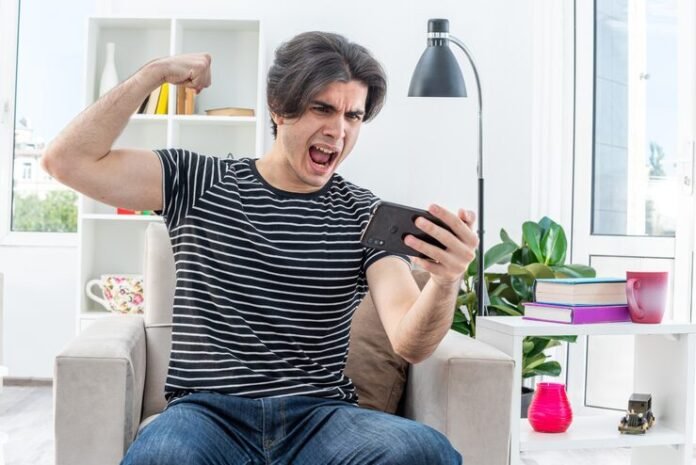 young-man-casual-clothes-playing-games-using-smartphone-happy-excited-raising-fist-like-winner-sitting-chair-light-living-room_141793-98896